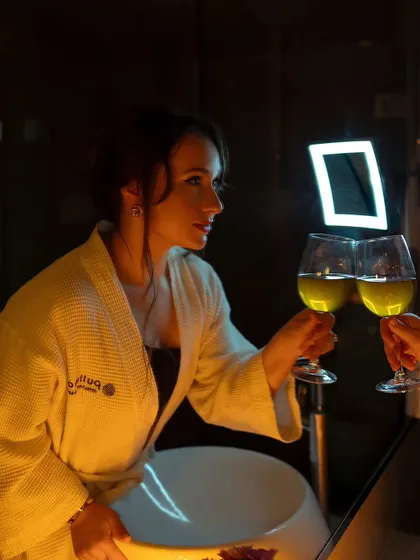 A low-light, intimate portrait of a woman in a bathrobe. The warm, candle-like lighting and the act of clinking glasses create a romantic and cozy atmosphere.