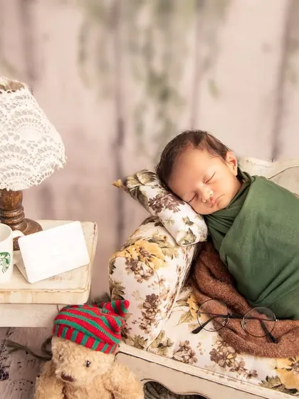 A cozy and relaxed scene with a newborn sleeping on a miniature floral sofa. The tiny coffee cup and glasses add a touch of whimsical storytelling.
