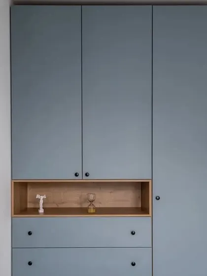 A modern, floor-to-ceiling wardrobe in a matte dusty blue finish. The design incorporates an open wooden niche and drawers, breaking the solid block of color and adding functional display space.