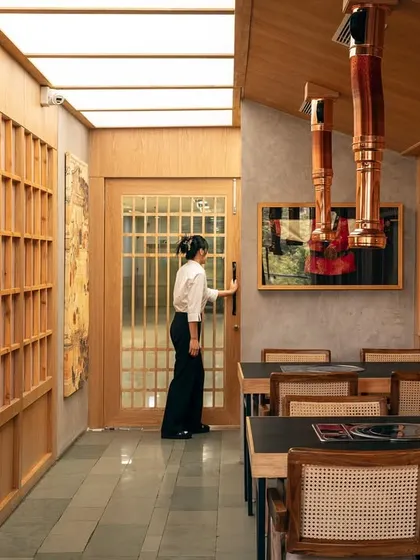 A staff member moves through the space, highlighting the functional flow of the design. The wooden lattice screens and paneled doors create a sense of rhythm and order, guiding movement through the restaurant.