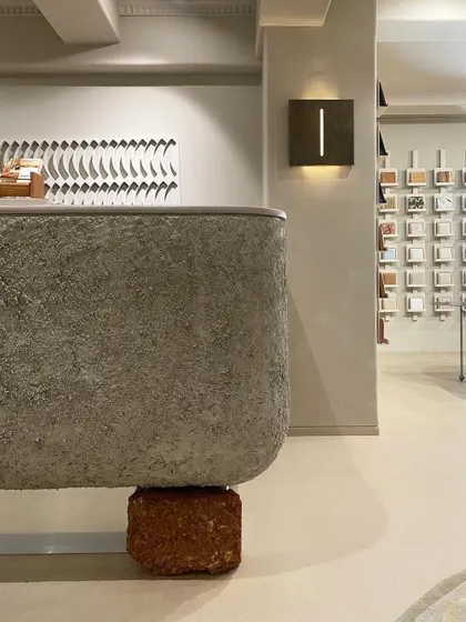 The reception desk is a statement piece, designed with a heavily textured concrete finish and balanced on a raw brick block. This juxtaposition of rough, industrial materials with the polished showroom floor exemplifies our approach of finding beauty in contrast.