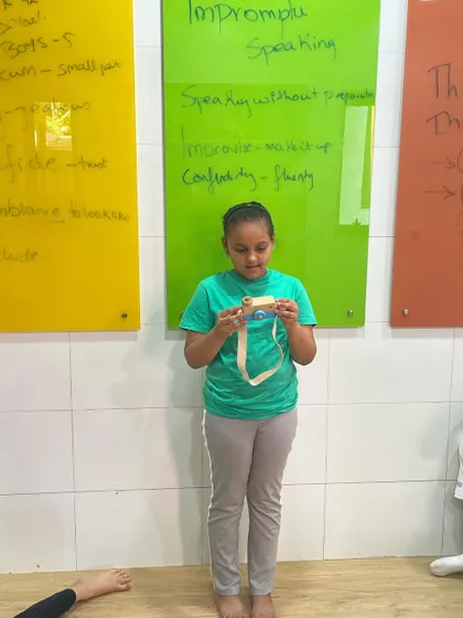 A student uses a toy camera as her prompt for an impromptu speaking exercise. This activity is designed to improve improvisation skills and the ability to structure a coherent narrative quickly.