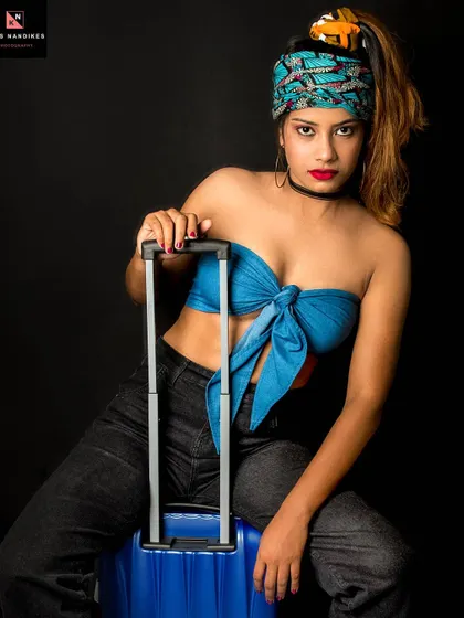 A powerful shot from the 'Caribbean Tourist' series. The model sits on a blue suitcase against a black background, her direct gaze and bold styling creating a striking image.