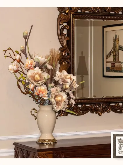 This beautifully arranged artificial floral design in a classic cream-colored vase is perfectly complemented by an ornate mirror, creating a vintage and elegant atmosphere.