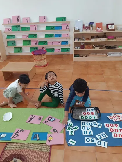 Two children work side-by-side on their respective mats. One explores language with sandpaper letters, while the other works on math with number cards, demonstrating parallel, focused learning.