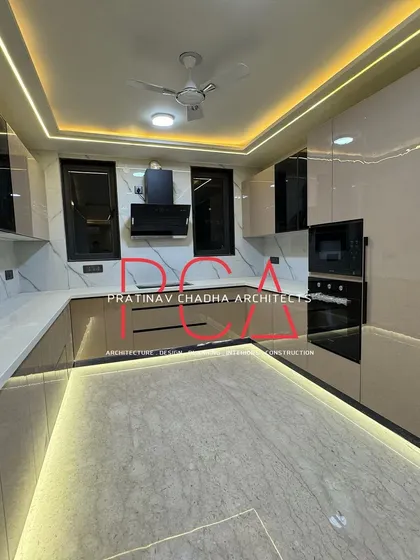 A modern, U-shaped modular kitchen from the Vasant Kunj luxury residence, designed for both functionality and aesthetic appeal. The space features high-gloss beige cabinetry, a marble-patterned backsplash, and integrated appliances, all illuminated by ambient ceiling and under-cabinet lighting.