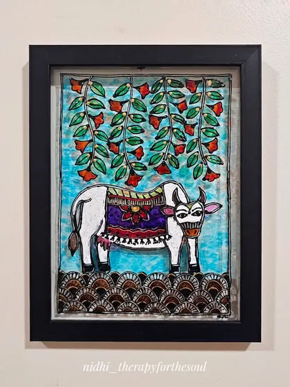 This Madhubani-style cow is painted on glass and framed. The artwork features a decorated cow under hanging flowers, with a patterned border at the base, all typical of the Madhubani aesthetic.