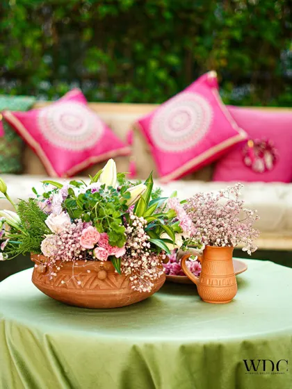 A tabletop detail from the 'Serene Sage' event. A terracotta bowl filled with pink and white flowers sits on a sage green tablecloth, with vibrant pink cushions in the background.