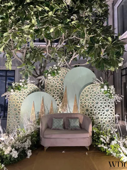 A serene and elegant stage setup. A plush sofa is set against a backdrop of mint green jali panels and surrounded by white flowers, creating a look that is both modern and timeless.