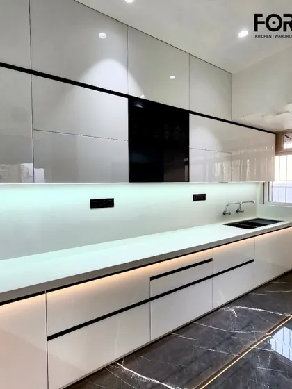 This kitchen features high-gloss white lacquered cabinets with a black handleless profile. The continuous LED lighting under the upper cabinets illuminates the entire workspace and enhances the reflective quality of the surfaces.