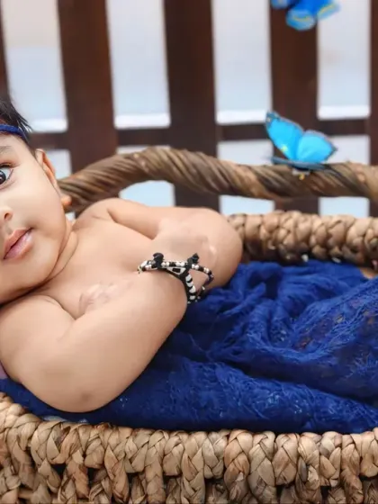 A beautiful baby with captivating eyes rests in a wicker basket, wrapped in a blue blanket and surrounded by butterflies.