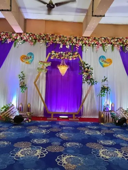 A unique engagement stage featuring a golden hexagonal frame with a small chandelier inside. The backdrop is decorated with rich purple and white drapes, accented with floral arrangements, perfect for a hall celebration.