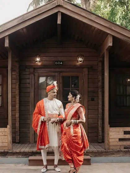 A full-length shot of the couple in their regal Maharashtrian wedding attire, standing before a charming wooden lodge.