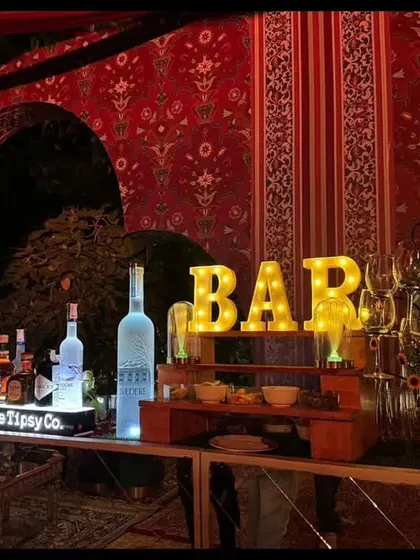 This bar setup for a wedding features a stunning backdrop of traditional Indian patterns in red. The illuminated 'BAR' sign and premium spirits on display create a warm and inviting focal point.