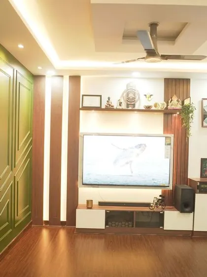 A full view of a living room renovation, featuring a rich green accent wall with decorative molding, a custom pooja cabinet, and a modern TV unit, all tied together with warm wooden flooring.