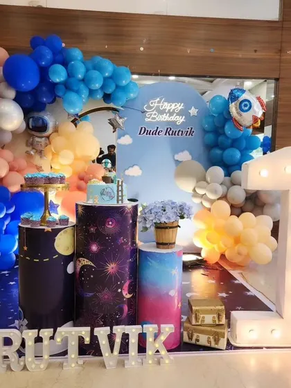 A cosmic adventure for Rutvik's first birthday with this space theme. We used a galaxy-print floor mat, planet-themed pedestals, and a blue and orange balloon garland with astronaut and rocket props.