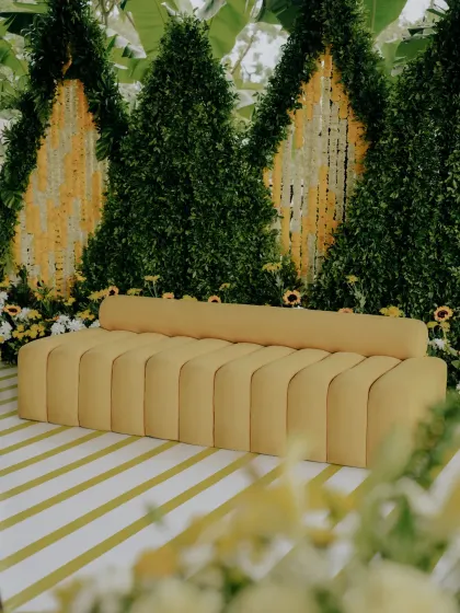A modern Haldi seating arrangement with a sleek yellow sofa set against a backdrop of green foliage and hanging yellow and white floral strings.