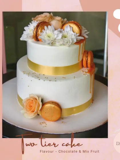A chic two-tier cake featuring a white and gold theme. This design is elevated with a gold ribbon, macarons, and fresh flowers, perfect for an elegant celebration.