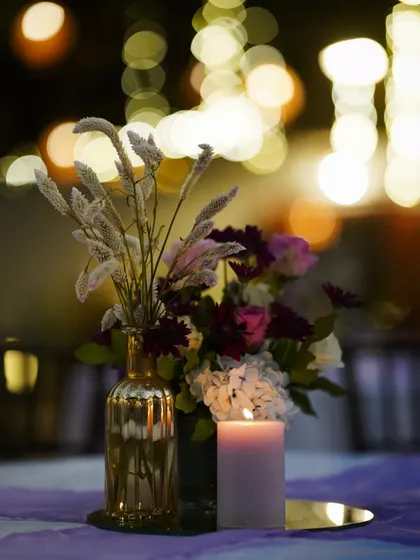 A simple yet elegant detail for a cocktail evening. A single candle and a delicate floral arrangement with dried grasses create a warm and inviting ambiance, beautifully captured against a backdrop of soft bokeh lights.