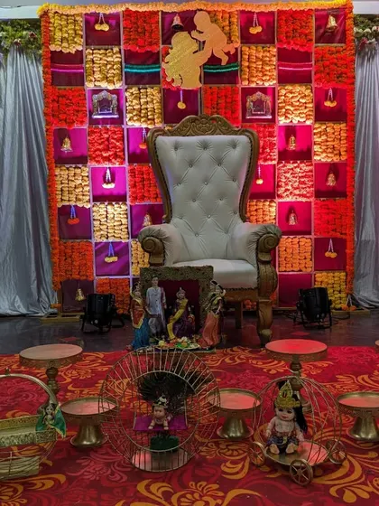 A complete Seemantham stage with a red and yellow floral grid backdrop. The setup includes a white throne, low stools, and various decorative items like miniature carriages and baskets.