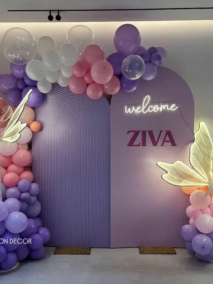 A chic and modern welcome decoration for baby Ziva. This setup uses pastel arched backdrops, organic balloon arrangements, and custom neon signage, complemented by elegant light-up butterfly props.