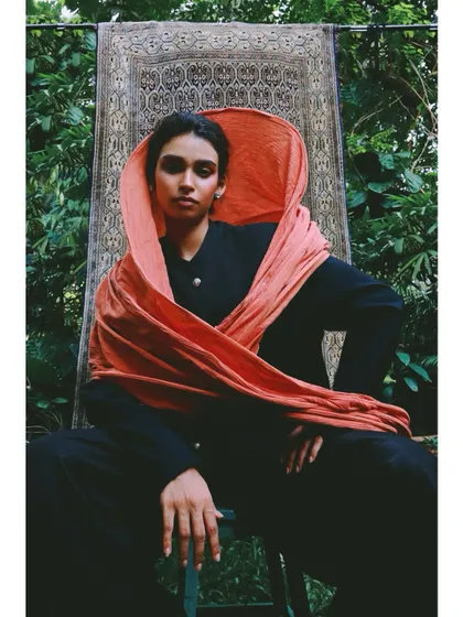 For this shot in the 'Roots' series, I styled a vibrant orange scarf as a hood against a simple black outfit. The tapestry in the background connects the outdoor setting to the indoor, cultural theme.
