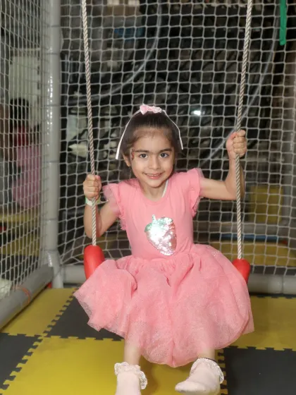 A little girl in a pink dress smiles brightly while enjoying the swing inside our netted play area.