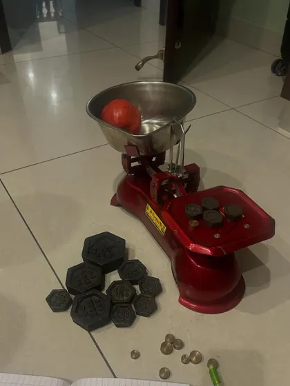 A real-world math lesson in our kitchen, using a traditional weighing scale with a tomato and weights. This shows how everyday objects can be used for experiential learning, teaching concepts like estimation, measurement, and calculation.