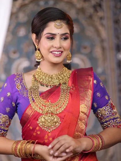 A classic South Indian bridal portrait. The bride is wearing a beautiful layered temple jewellery set that stands out against her red and purple silk saree.