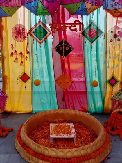 A vibrant Haldi ceremony space featuring a multi-colored backdrop with hanging kites and umbrellas. The 'urli' is filled with flower petals, ready for the rituals.