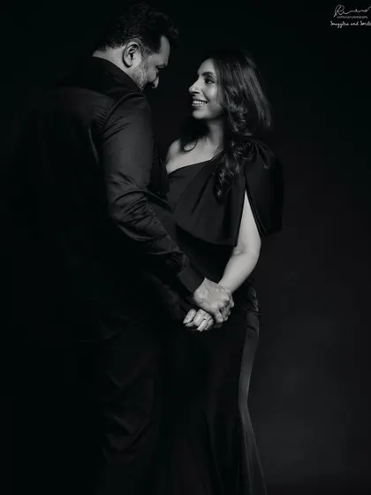 A full-length couple portrait that feels like a classic movie scene. The dark attire and deep shadows create a sense of timeless romance in this black and white photo.