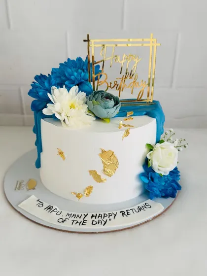 An elegant white cake with a royal blue drip, gold leaf accents, and a beautiful arrangement of blue and white flowers.