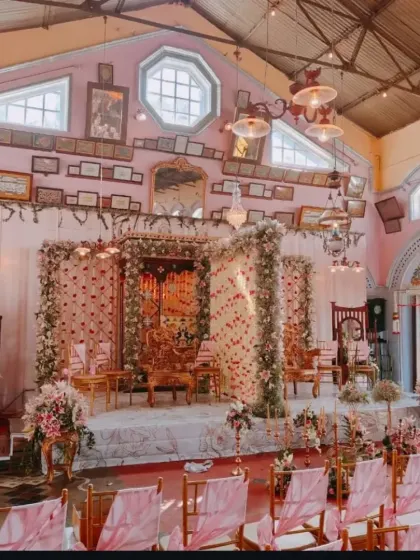 A stunning heritage hall in Ooty transformed for a wedding ceremony. The decor complements the venue's vintage charm with pastel pink drapes and lush floral arrangements on the stage.