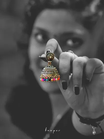 A hand holds up a colorful jhumka earring, which is the only element in color against a black and white background. This selective color technique makes the product the undeniable hero of the shot.