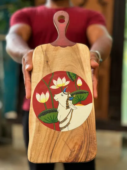 A hand-painted wooden platter featuring a Pichwai-style cow amidst lotuses. These organically shaped platters are perfect as festive decor, either hung on a wall or used as a centerpiece.