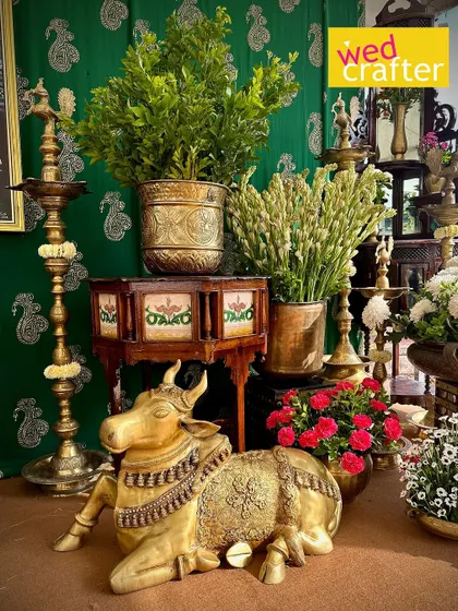 A seated brass Nandi statue surrounded by traditional lamps, wooden furniture, and lush greenery, creating a sacred and beautiful corner.