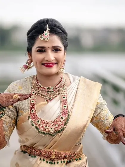 What a happy bride. For her wedding, we chose a gorgeous Kundan and gemstone bridal set that perfectly matched her off-white and red saree. Her smile makes the look even more beautiful.