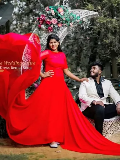 A couple poses with a decorative moon prop. The vibrant red trail gown adds a bold splash of color to the whimsical garden setting.