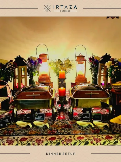 A rustic and romantic dinner setup. The table is covered with a traditional patterned cloth and lit by lanterns and candles, creating a warm and inviting atmosphere for an intimate meal.