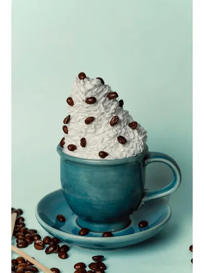 A creative and playful shot of a coffee cup overflowing with whipped cream and coffee beans. The pastel blue background and matching cup create a fun, stylized image perfect for social media marketing.