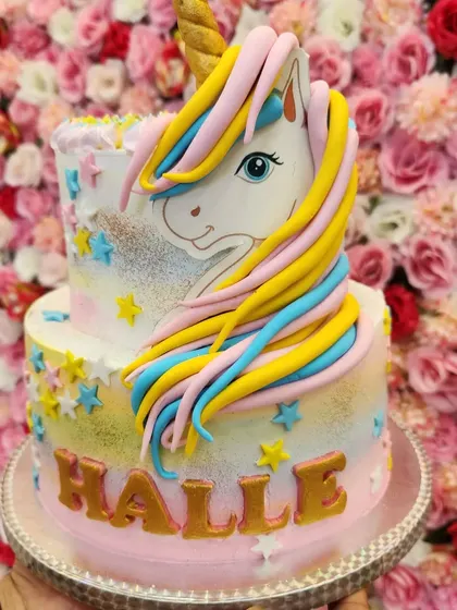 A dreamy two-tiered unicorn cake with a pastel rainbow airbrushed effect. The hand-sculpted fondant unicorn and golden name make it a personalized fantasy creation.