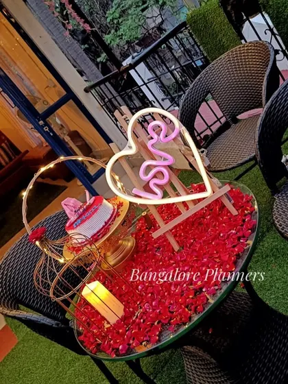 A sweet and simple table setup for two. This romantic arrangement on a cafe terrace features a table covered in red rose petals, a small cake, and a heart-shaped "LOVE" neon sign.