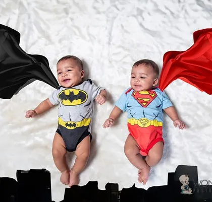 Double the trouble, double the fun! These twin baby boys are dressed as little superheroes, Batman and Superman, complete with capes that look like they're flying over a city skyline.