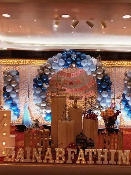 A grand stage setup for a Frozen-themed 4th birthday party in a hall. This decoration includes a large balloon arch in blue and silver, character standees of Elsa and Olaf, and a full display with marquee numbers and letters.