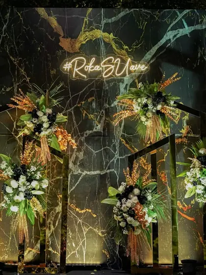A different take on a photo backdrop, featuring a black marble wall with a neon sign. Geometric stands hold arrangements of black, white, and gold florals, creating a chic and modern art piece.