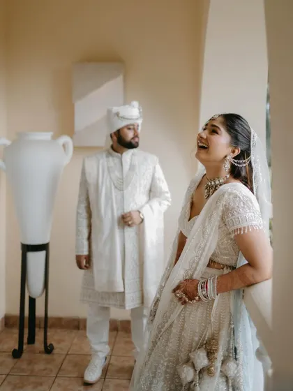 A candid moment of laughter between the couple on a balcony, capturing their genuine joy and easy chemistry in their all-white wedding outfits.