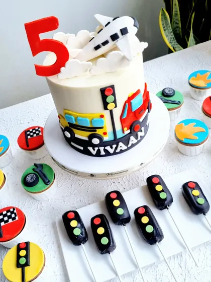 A vehicle-themed cake with matching cupcakes and cakesicles. The design includes a bus, truck, and airplane, with traffic light cakesicles.