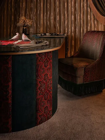 The curved client table is clad in alternating panels of rich red and deep teal fabric, creating a bold statement. The custom-embroidered swivel chair adds another layer of texture and craft, completing this maximalist composition.