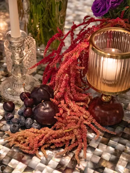 An artistic detail from the tablescape, featuring red amaranthus, fresh plums, and grapes alongside a ribbed glass candle holder. We often incorporate fruits and other natural elements to add texture and a touch of the unexpected.