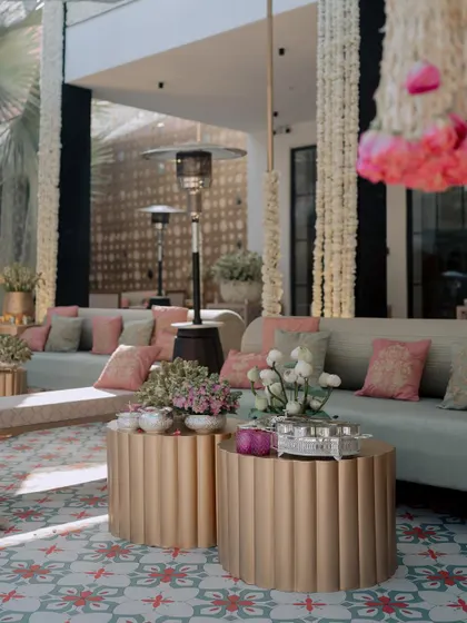 A lounge area with modern gold coffee tables and soft, comfortable seating. The space is decorated with hanging floral strings and traditional Indian textiles.